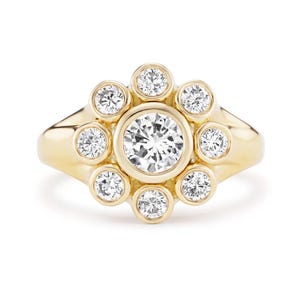 May include: A gold ring featuring a central, large, round, clear gemstone surrounded by smaller, clear gemstones, creating a floral or sunburst design. The ring has a smooth, polished band.