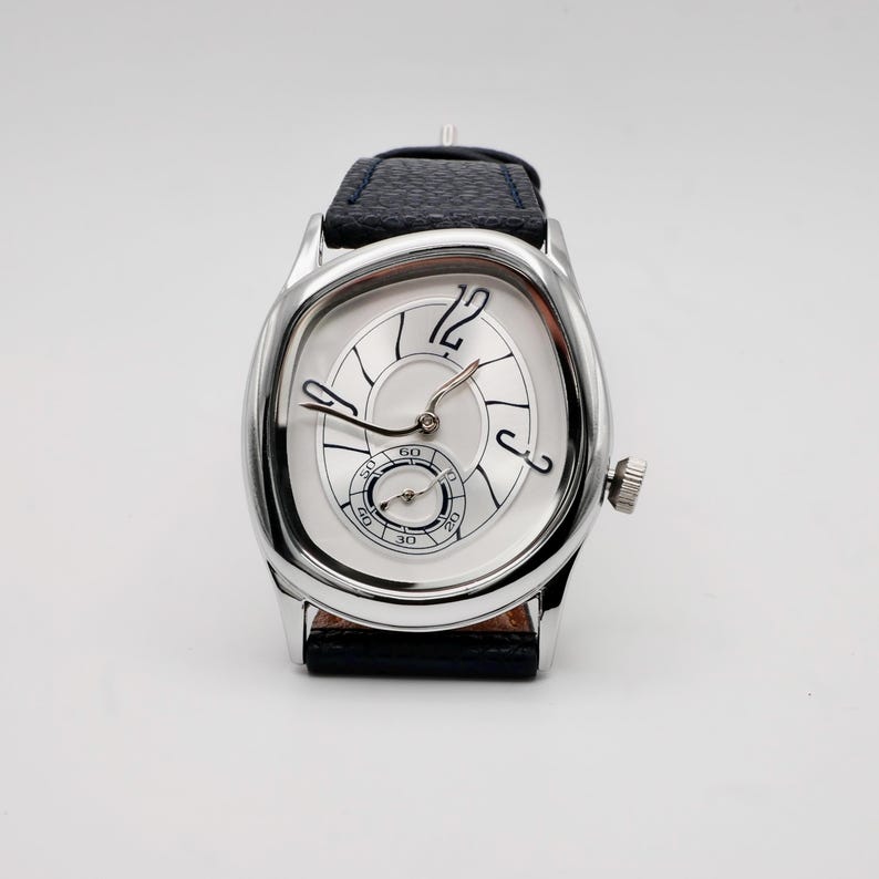 May include: A silver-toned wristwatch with a unique oval-shaped face and a black leather band. The watch face is white with black numerals and hands, and a small subdial. The watch has a crown on the side.