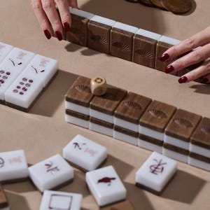 May include: Mahjong game set with brown wooden tiles featuring a fan design, white tiles with red symbols, and a wooden die. The game pieces are arranged on a light brown surface, with hands visible.