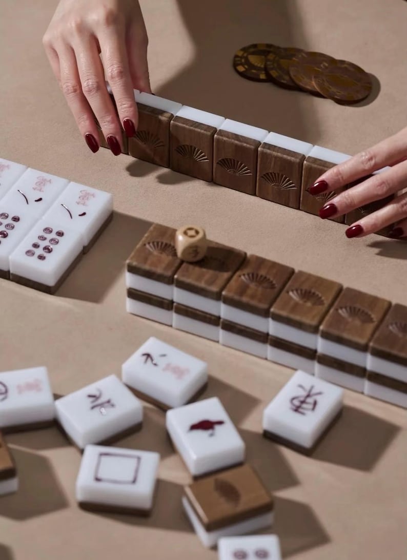 May include: Mahjong game set with brown wooden tiles featuring a fan design, white tiles with red symbols, and a wooden die. The game pieces are arranged on a light brown surface, with hands visible.