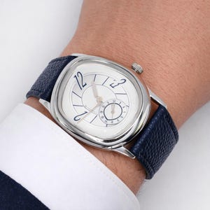May include: A silver wristwatch with a white face and blue Arabic numerals. The watch has a small seconds dial and a dark blue leather strap. The watch is on a wrist, with a white shirt cuff visible.