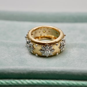 Puede incluir: Un anillo ancho, dorado, con una superficie texturizada. El anillo est&aacute; adornado con dise&ntilde;os florales con diamantes y peque&ntilde;os detalles plateados. El interior del anillo tambi&eacute;n es dorado, creando una apariencia lujosa. Una joya.