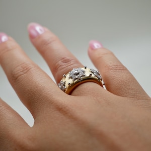 Puede incluir: Un anillo de oro con una banda ancha, adornado con m&uacute;ltiples grupos de diamantes en forma de flor y peque&ntilde;os detalles plateados en relieve. El anillo est&aacute; en un dedo, mostrando su intrincado dise&ntilde;o.