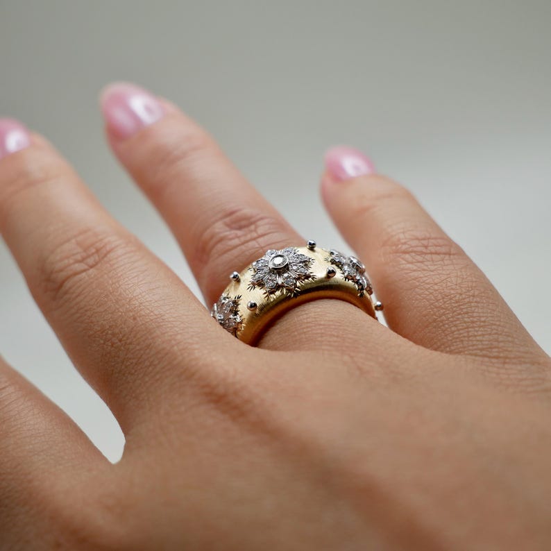 Puede incluir: Un anillo de oro con una banda ancha, adornado con m&uacute;ltiples grupos de diamantes en forma de flor y peque&ntilde;os detalles plateados en relieve. El anillo est&aacute; en un dedo, mostrando su intrincado dise&ntilde;o.