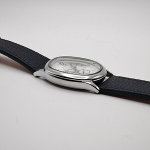 May include: A silver wristwatch with a dark blue leather band. The watch has a unique oval face with a silver dial and black numerals. The watch hands are black. The band is textured.
