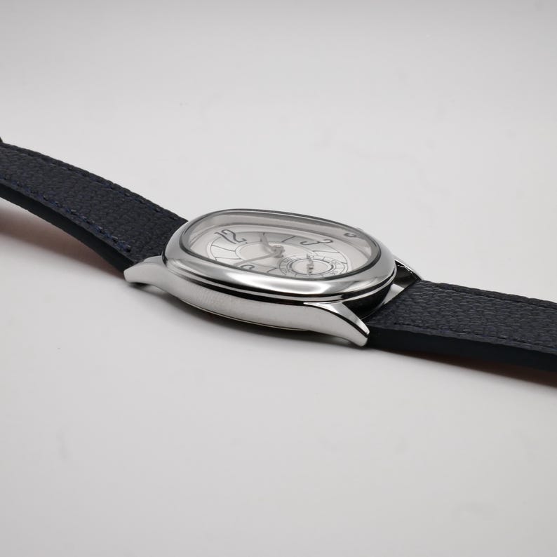 May include: A silver wristwatch with a dark blue leather band. The watch has a unique oval face with a silver dial and black numerals. The watch hands are black. The band is textured.