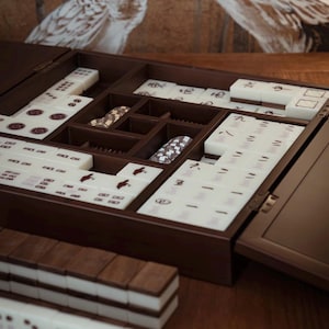 May include: A dark brown wooden Mahjong set with ivory tiles. The set is open, showing compartments for tiles and accessories. The tiles display Chinese characters and symbols. The set is on a wooden surface.