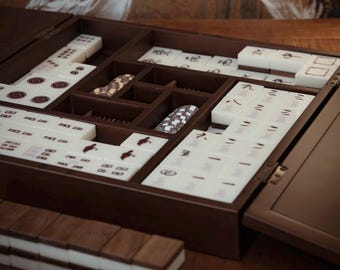 May include: A dark brown wooden Mahjong set with ivory-colored tiles. The set is open, revealing organized compartments for tiles and accessories. The tiles feature Chinese characters and symbols. The set is on a wooden surface.