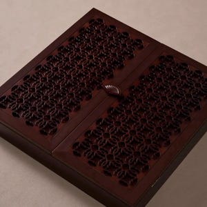 May include: A dark brown wooden box with a decorative floral pattern carved into the lid. The box has a rectangular shape and a small, fan-shaped handle. The wood has a rich, warm tone.