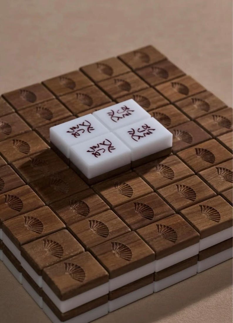 May include: A set of wooden Mahjong tiles with a fan design carved into the wood. Four white tiles with red characters are stacked on top of the wooden tiles. The tiles are arranged in a grid pattern.
