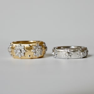 Puede incluir: Se muestran dos anillos de banda ancha, uno dorado y otro plateado. Ambos anillos presentan un dise&ntilde;o floral con piedras preciosas transparentes. El anillo dorado tiene un acabado cepillado, mientras que el anillo plateado tiene un acabado pulido.