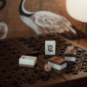 May include: A close-up of a dark brown, carved wooden table with several Mahjong tiles and dice. The tiles are white with black characters. The dice are light brown. A crane painting is in the background.