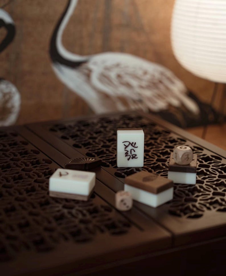 May include: A close-up of a dark brown, carved wooden table with several Mahjong tiles and dice. The tiles are white with black characters. The dice are light brown. A crane painting is in the background.