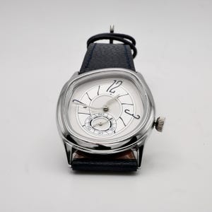 May include: A silver-toned wristwatch with a square face and a white dial. The watch features black hands, a small seconds dial, and a dark blue leather band. The watch has a crown on the side.