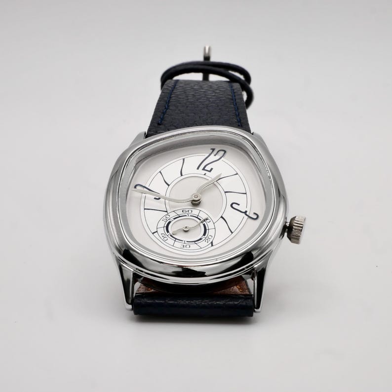 May include: A silver-toned wristwatch with a square face and a white dial. The watch features black hands, a small seconds dial, and a dark blue leather band. The watch has a crown on the side.