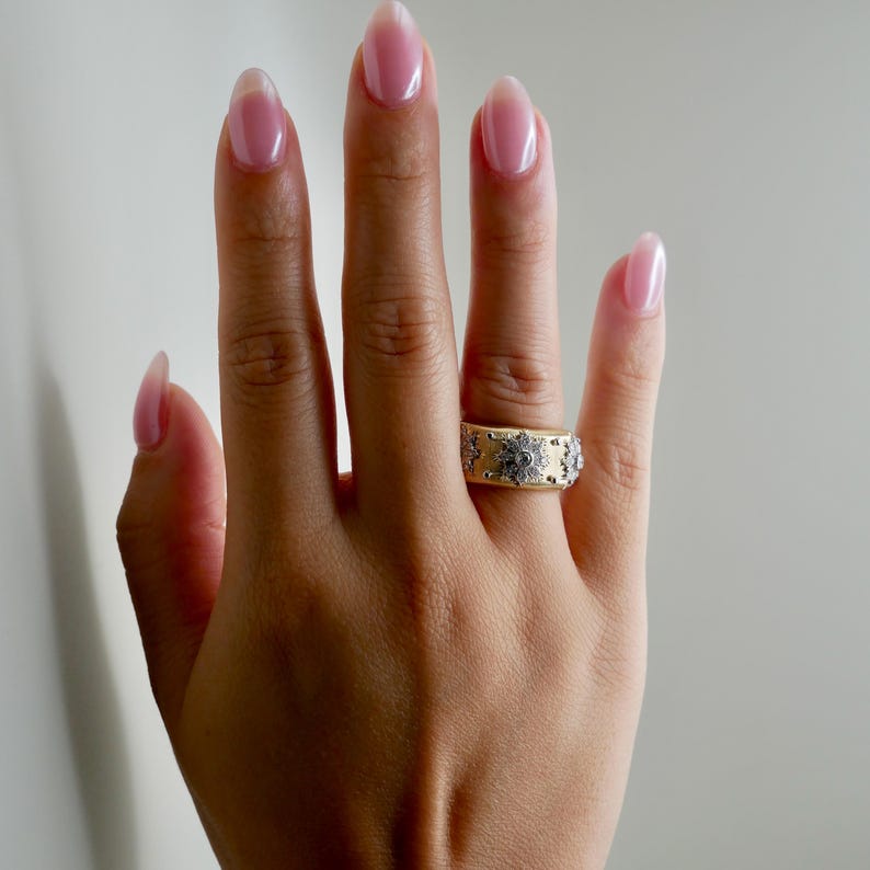 Puede incluir: Un anillo dorado con una banda ancha, adornado con un patr&oacute;n floral repetido y detalles plateados. El anillo se muestra en una mano con u&ntilde;as cuidadas, destacando el dise&ntilde;o de la joya. El dise&ntilde;o del anillo es intrincado y detallado.