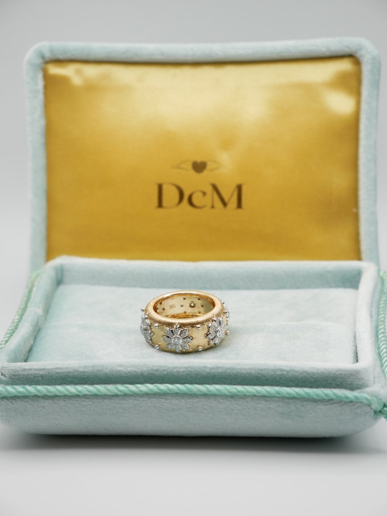 Puede incluir: Un anillo de oro con dise&ntilde;o floral, engastado con diamantes, descansa en un joyero de terciopelo azul claro. La caja tiene un interior dorado y se ven las iniciales "DcM".
