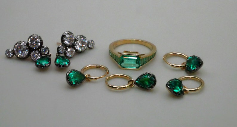 May include: A set of emerald green and diamond jewelry featuring a ring with a large emerald green stone, a pair of earrings with emerald green stones, and a pair of hoop earrings with emerald green stones.