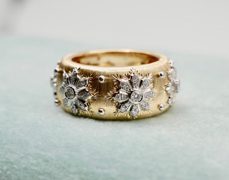 Puede incluir: Un anillo ancho de oro con una superficie texturizada, adornado con dise&ntilde;os en forma de copo de nieve. Cada copo de nieve presenta un diamante central y piedras m&aacute;s peque&ntilde;as. Peque&ntilde;as cuentas plateadas est&aacute;n intercaladas alrededor del anillo.