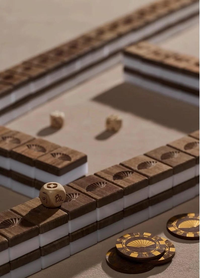 May include: A wooden Mahjong set with brown tiles and white inserts. The tiles are arranged in a game layout, with dice and decorative chips visible. The tiles have a fan design carved into them. The game is set on a neutral surface.