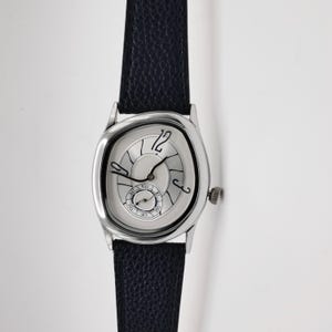 May include: A silver-toned wristwatch with a unique, distorted oval face and a black leather band. The watch features a small seconds dial and black hands against a light-colored face. The watch case is approximately 1.5 inches in length.