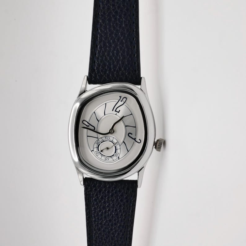 May include: A silver-toned wristwatch with a unique, distorted oval face and a black leather band. The watch features a small seconds dial and black hands against a light-colored face. The watch case is approximately 1.5 inches in length.