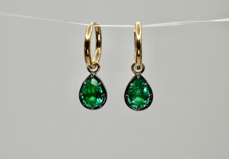 May include: A pair of gold hoop earrings with a teardrop-shaped emerald gemstone dangling from each hoop. The gemstones are set in a black metal bezel.