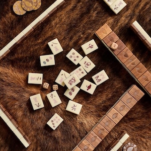 May include: A top-down view of a Mahjong set with ivory tiles, wooden racks, and brown game chips. The tiles have red symbols and numbers. The racks are brown with white accents. The game pieces are scattered on a brown, textured surface.