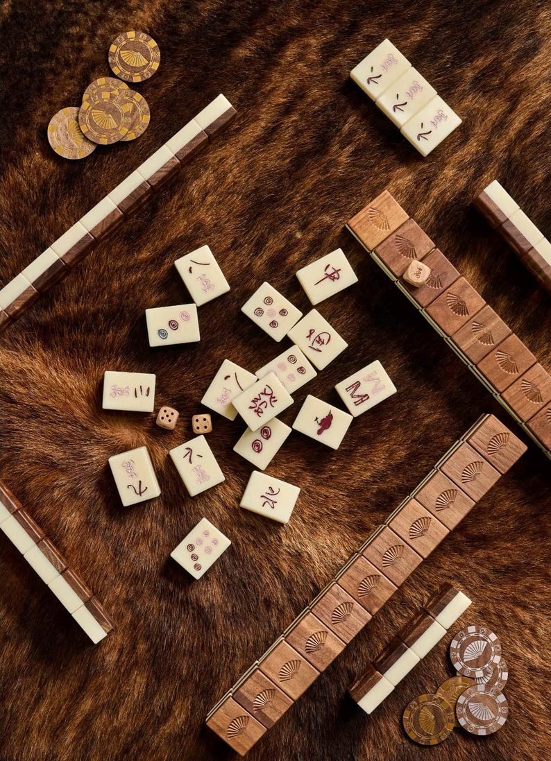 May include: A top-down view of a Mahjong set with ivory tiles, wooden racks, and brown game chips. The tiles have red symbols and numbers. The racks are brown with white accents. The game pieces are scattered on a brown, textured surface.