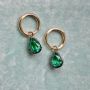 May include: A pair of gold hoop earrings with a teardrop-shaped green gemstone pendant hanging from each hoop.
