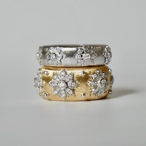 Puede incluir: Dos anillos apilados, uno plateado y otro dorado, cada uno adornado con patrones florales de gemas brillantes. Los anillos tienen una superficie texturizada y un dise&ntilde;o de banda ancha, mostrando una est&eacute;tica lujosa.