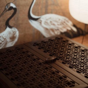 May include: A dark brown wooden cabinet with intricate, openwork floral patterns on its doors. The cabinet has a small, decorative handle. In the background, a wall features a painting of two white and black cranes.