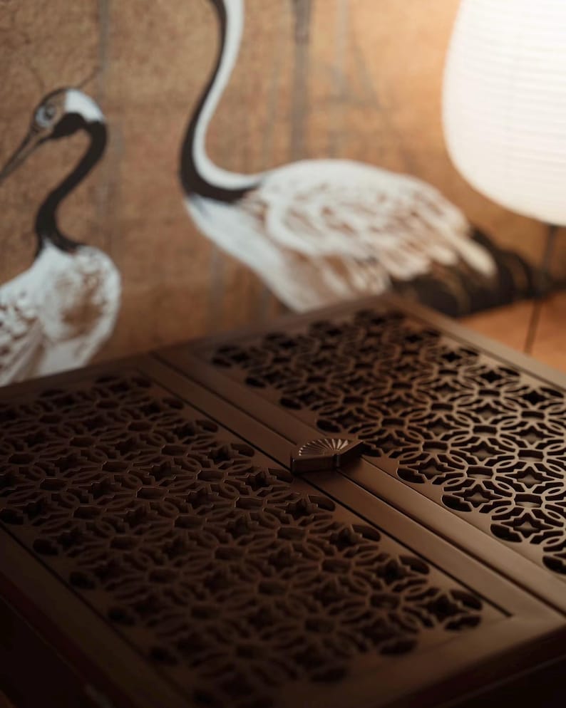 May include: A dark brown wooden cabinet with intricate, openwork floral patterns on its doors. The cabinet has a small, decorative handle. In the background, a wall features a painting of two white and black cranes.