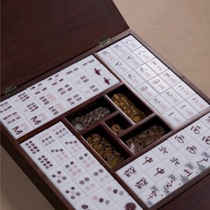 May include: A wooden Mahjong set with a dark brown finish. The open box reveals tiles with red and black symbols, along with wooden and decorative pieces. The tiles are arranged in compartments within the box.