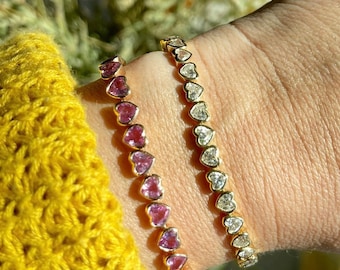 May include: Two gold heart-shaped bracelets with pink and clear gemstones. The bracelets are worn on a person's wrist.