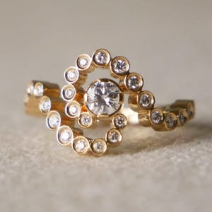 May include: A gold ring featuring a large central diamond encircled by smaller diamonds. The band curves, with more small diamonds set along the sides. The ring is set against a neutral background. The ring's design is elegant.