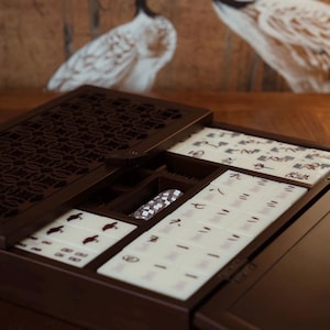 May include: A dark brown wooden Mahjong set with open compartments revealing tiles with Chinese characters and symbols. The set includes a patterned dice holder and is displayed on a wooden surface. The background features a painting of two cranes.