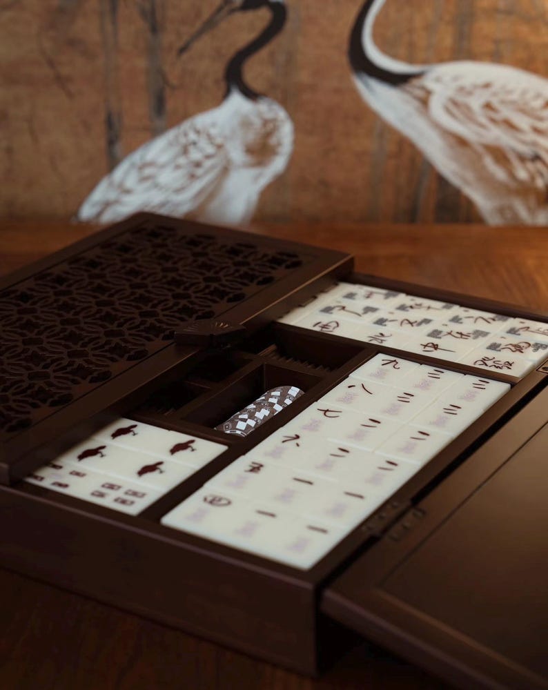 May include: A dark brown wooden Mahjong set with open compartments revealing tiles with Chinese characters and symbols. The set includes a patterned dice holder and is displayed on a wooden surface. The background features a painting of two cranes.
