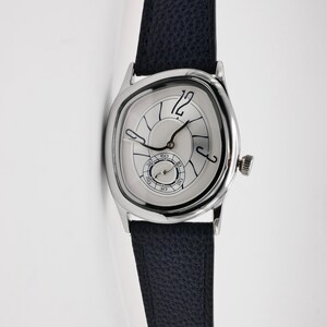 May include: A silver wristwatch with a unique, asymmetrical face and a dark blue leather band. The watch face features black numerals and hands, with a small seconds dial. The watch has a textured crown and a classic design.