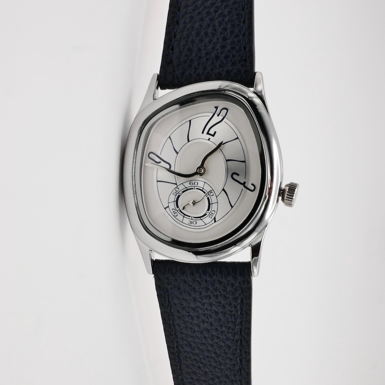 May include: A silver wristwatch with a unique, asymmetrical face and a dark blue leather band. The watch face features black numerals and hands, with a small seconds dial. The watch has a textured crown and a classic design.