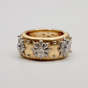 Puede incluir: Un anillo ancho de oro con acabado cepillado, adornado con dise&ntilde;os en forma de copo de nieve incrustados con diamantes. Peque&ntilde;as cuentas plateadas est&aacute;n espaciadas uniformemente alrededor de los bordes. El anillo se presenta sobre un fondo blanco.