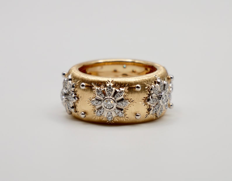 Puede incluir: Un anillo ancho de oro con acabado cepillado, adornado con dise&ntilde;os en forma de copo de nieve incrustados con diamantes. Peque&ntilde;as cuentas plateadas est&aacute;n espaciadas uniformemente alrededor de los bordes. El anillo se presenta sobre un fondo blanco.