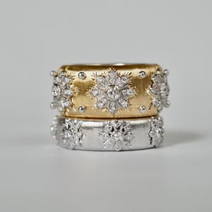 Puede incluir: Dos anillos apilados, uno dorado y otro plateado, cada uno adornado con intrincados dise&ntilde;os de diamantes en forma de copo de nieve. El anillo dorado tiene un acabado texturizado y peque&ntilde;os detalles redondos. El anillo plateado tiene un acabado liso.