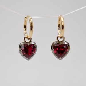 May include: A pair of gold hoop earrings with a dangling red heart charm. The heart charm has a silver border and a red gemstone in the center.