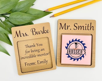 May include: Two wooden desk organizers with personalized text. One reads "Mrs. Burke" with a message, the other "Mr. Smith" with a pink sticky note and a logo. Two pencils and green leaves are in the background.