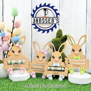 May include: Three wooden bunny money holders with personalized names: Haylie, Samantha, and Jack. Each bunny is holding a rolled-up dollar bill. The bunnies are painted in pastel colors and have a bow or a tie. The sign at the bottom reads "Money Not Included".