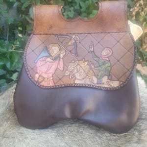 May include: A brown leather pouch featuring a flap with an embossed scene of a person aiming a crossbow at a horse and rider. The leather has a diamond-patterned background and scrollwork details. The lower section of the pouch is smooth dark brown.
