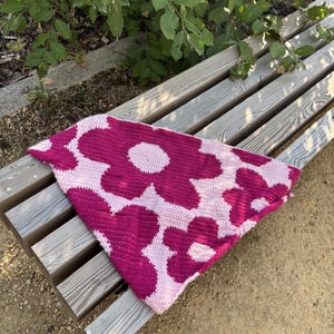 May include: A triangular crochet bandana featuring a repeating pattern of large, bold magenta flowers on a light pink background. The textile accessory is crafted with a dense stitch pattern, creating a flat, textured surface with distinct floral shapes.