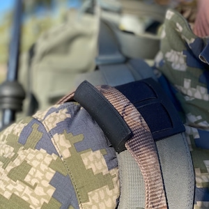 Puede incluir: Primer plano de una correa de hombro acolchada negra para una mochila. La correa está unida a una mochila de camuflaje marrón y gris.