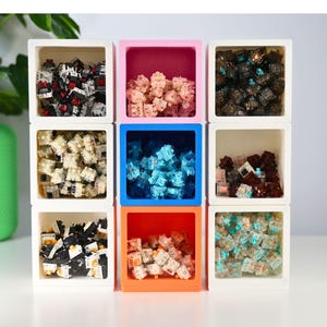 May include: Nine square containers, each filled with different colored mechanical keyboard switches. The containers are white, pink, blue, and orange. The switches are various colors, including red, black, and clear.